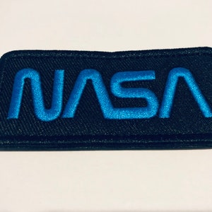 Op de afbeelding: Een zwarte geborduurde patch met het woord "NASA" in blauwe letters.
