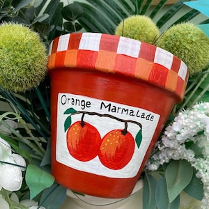 May include: A hand-painted orange flower pot with a white label that reads "Orange Marmalade." The pot has a checkered rim and a painted illustration of two oranges. The pot is surrounded by greenery and white flowers.