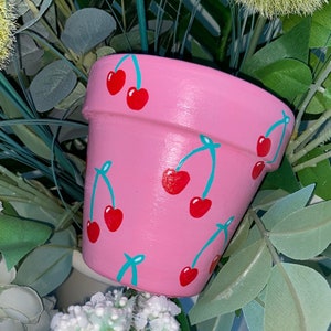 May include: A pink ceramic planter with a cherry design. The planter has a smooth, matte finish and is decorated with red cherries and green stems.