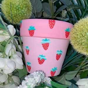 May include: A pink terracotta flower pot with a strawberry pattern. The pot is painted with red and green strawberries on a light pink background.