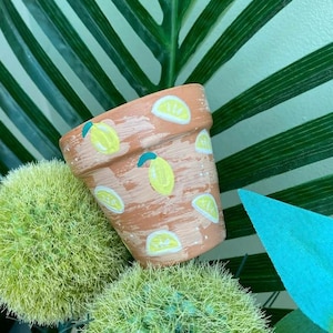 May include: A terracotta flower pot painted with a lemon pattern. The pot is decorated with yellow lemons and green leaves.