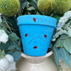 May include: A sky blue ceramic plant pot adorned with red ladybugs and a dotted flight path. The pot is set against a backdrop of green foliage and white flowers, creating a cheerful, garden-themed aesthetic.