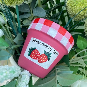 May include: A pink ceramic flower pot with a strawberry jam design. The pot has a red and white chequered rim and a label with the words "Strawberry Jam" and a graphic of two strawberries. The pot is surrounded by greenery.