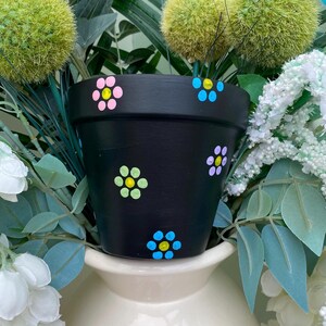 May include: A black painted plant pot decorated with colourful, hand-painted flowers. The flowers are in shades of pink, blue, yellow, and green. The pot is filled with greenery and white flowers, and sits on a cream-coloured vase.