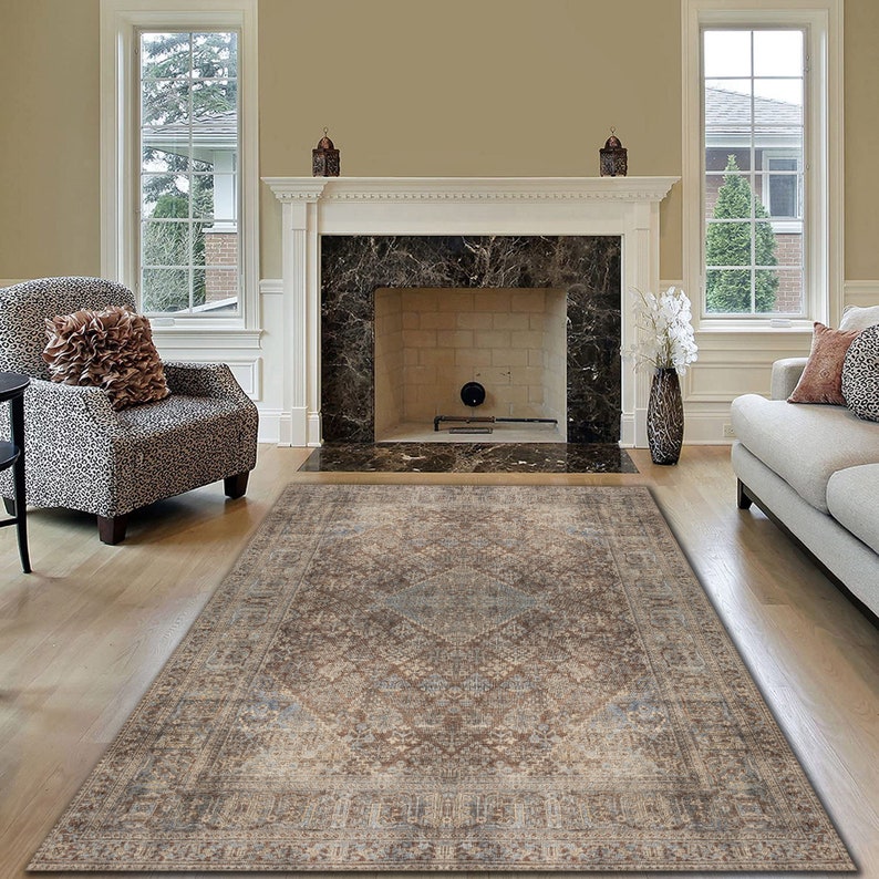 May include: A brown and beige area rug with a traditional Persian design. The rug is laid out on a hardwood floor in a living room with a fireplace and furniture.