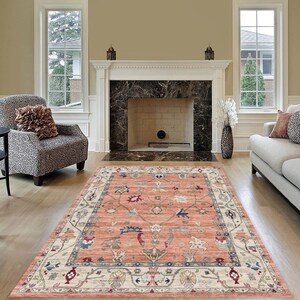 May include: A large, intricately patterned area rug with a cream and peach color scheme. The rug features a central medallion with floral and geometric designs. The rug is laid out in a living room setting with a fireplace and furniture.