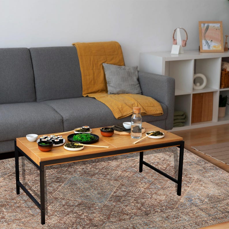 May include: A wooden coffee table with a black metal frame. The table is set with a variety of snacks, including sushi rolls, seaweed salad, and chopsticks. The table is in front of a gray sofa with a yellow throw blanket.