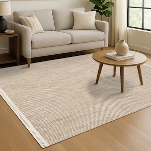 Puede incluir: Una alfombra beige tejida con un borde blanco, colocada sobre un suelo de madera. La alfombra rectangular tiene un patrón texturizado. Un sofá de color claro, una mesa de centro de madera y una mesa auxiliar están en el fondo.