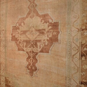 May include: A large, rectangular rug with a central medallion design in shades of rust and beige. The rug has an aged, vintage appearance, with a faded color palette and intricate border details. The rug is displayed against a white brick wall.