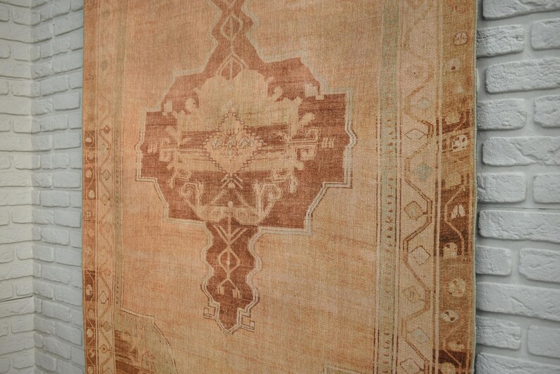May include: A large, rectangular rug with a central medallion design in shades of rust and beige. The rug has an aged, vintage appearance, with a faded color palette and intricate border details. The rug is displayed against a white brick wall.