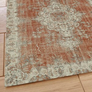 Puede incluir: Una alfombra de estilo vintage con un aspecto desgastado y descolorido. La alfombra presenta un dise&ntilde;o central de medall&oacute;n en color beige claro con detalles intrincados. El fondo de la alfombra es de un color naranja oxidado c&aacute;lido con un patr&oacute;n sutil.