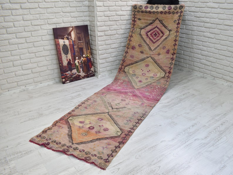 K&ouml;nnte beinhalten: Ein langer, rechteckiger Teppich mit einem geometrischen Muster in Rosa, Beige und Wei&szlig;. Der Teppich hat einen Fransenrand und ist aus einem weichen, gewebten Material gefertigt.