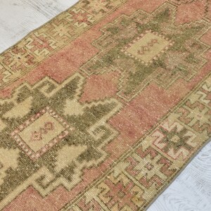Puede incluir: Una alfombra turca vintage de corredor con fondo rosa y beige. La alfombra tiene un patr&oacute;n geom&eacute;trico con un medall&oacute;n central en tonos verdes y beige.