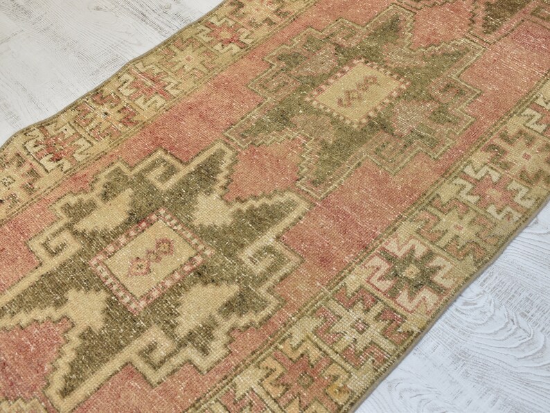 Puede incluir: Una alfombra turca vintage de corredor con fondo rosa y beige. La alfombra tiene un patr&oacute;n geom&eacute;trico con un medall&oacute;n central en tonos verdes y beige.