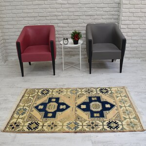 May include: A beige rug with a blue and cream geometric pattern. The rug is placed on a white wood floor in front of two chairs, one red and one gray.