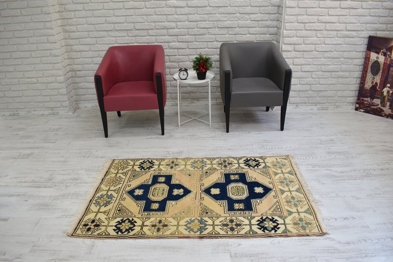 May include: A beige rug with a blue and cream geometric pattern. The rug is placed on a white wood floor in front of two chairs, one red and one gray.