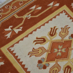 Puede incluir: Un primer plano de una alfombra turca tradicional con fondo blanco y patrones intrincados en tonos naranja, amarillo y marr&oacute;n. La alfombra presenta una forma de diamante central con motivos florales estilizados y dise&ntilde;os geom&eacute;tricos.