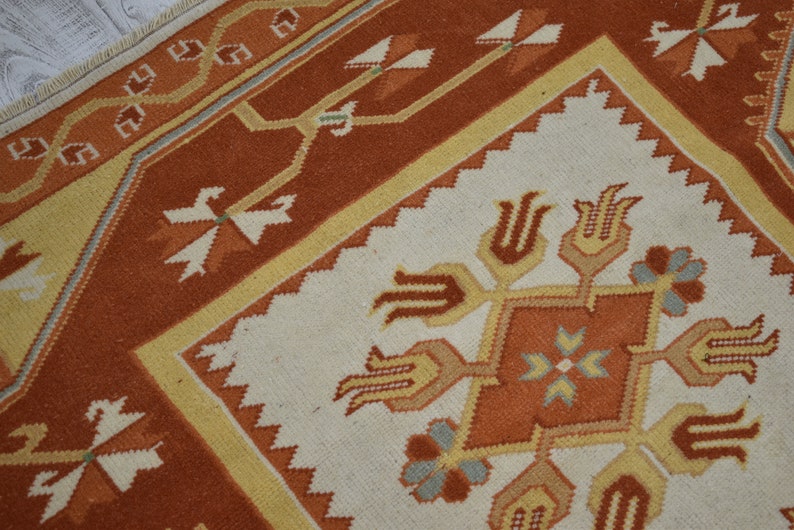 Puede incluir: Un primer plano de una alfombra turca tradicional con fondo blanco y patrones intrincados en tonos naranja, amarillo y marr&oacute;n. La alfombra presenta una forma de diamante central con motivos florales estilizados y dise&ntilde;os geom&eacute;tricos.