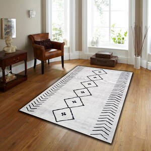 May include: A white rug with a black geometric pattern. The pattern features a series of black squares with a cross shape inside, arranged in a diagonal line. The rug has a chevron border.