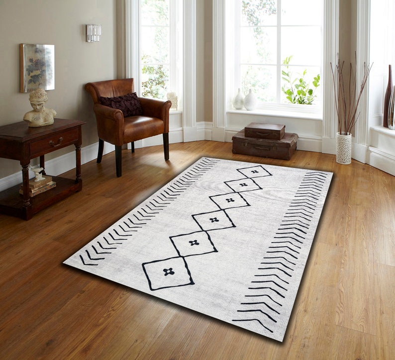 May include: A white rug with a black geometric pattern. The pattern features a series of black squares with a cross shape inside, arranged in a diagonal line. The rug has a chevron border.