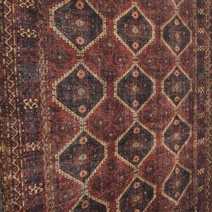 Op de afbeelding: Een vintage vloerkleed met een geometrisch patroon. Het vloerkleed heeft een herhalend ruitontwerp in tinten rood, zwart en beige. Het vloerkleed wordt gepresenteerd tegen een witte bakstenen muur.