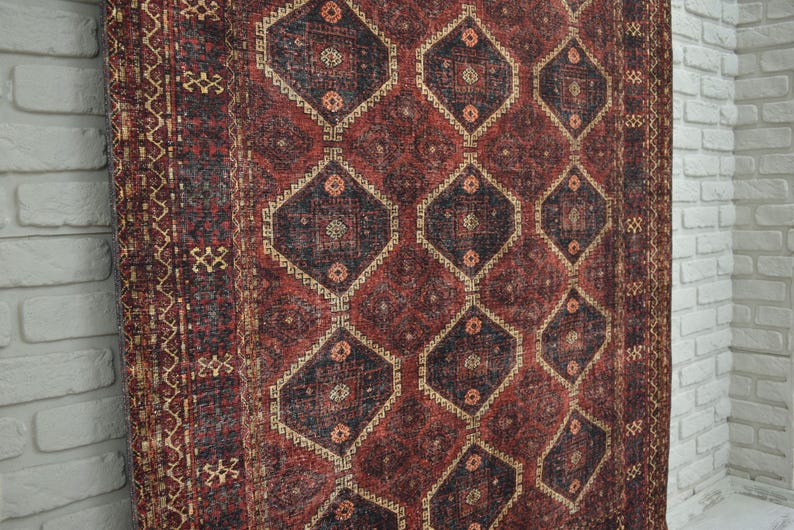 Op de afbeelding: Een vintage vloerkleed met een geometrisch patroon. Het vloerkleed heeft een herhalend ruitontwerp in tinten rood, zwart en beige. Het vloerkleed wordt gepresenteerd tegen een witte bakstenen muur.