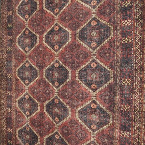 Op de afbeelding: Een gedetailleerde foto van een vintage tapijt met een geometrisch patroon. Het tapijt heeft een dieprood en marineblauw kleurenschema met ruit- en bloemmotieven. De randen hebben een decoratieve rand.