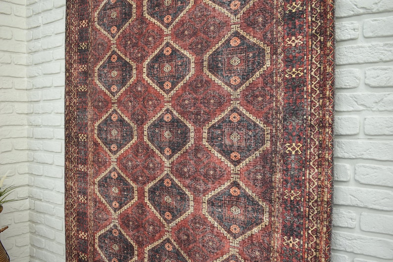 Op de afbeelding: Een gedetailleerde foto van een vintage tapijt met een geometrisch patroon. Het tapijt heeft een dieprood en marineblauw kleurenschema met ruit- en bloemmotieven. De randen hebben een decoratieve rand.