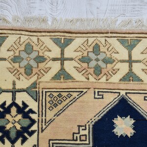 May include: A close-up of a beige rug with a blue and green geometric pattern. The rug has a fringe border.