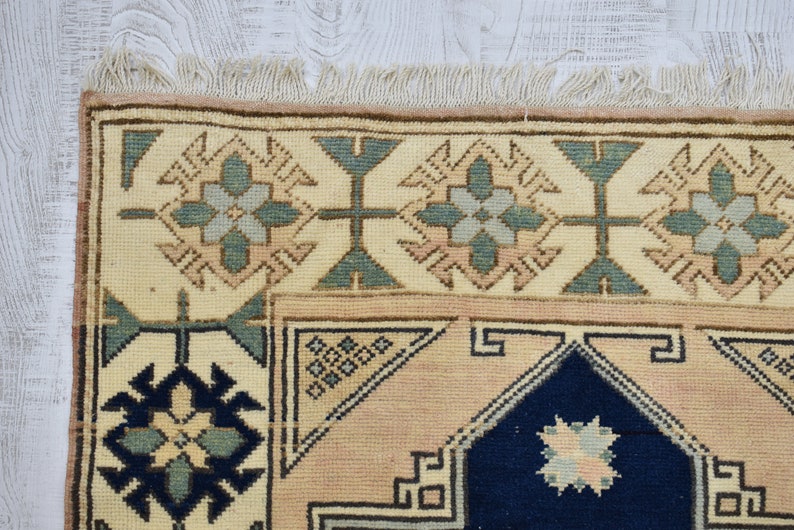 May include: A close-up of a beige rug with a blue and green geometric pattern. The rug has a fringe border.