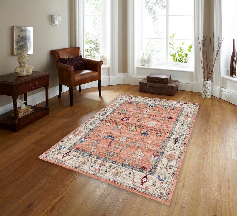 May include: A pink and cream area rug with a floral pattern. The rug has a detailed border with a repeating design. The rug is laid out on a hardwood floor in a living room setting.