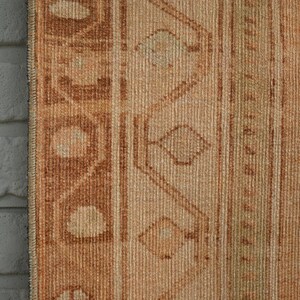 May include: Close-up of a section of a vintage rug with a brick wall on the left. The rug features a warm color palette of beige, brown, and tan. Geometric patterns and vertical stripes are visible.