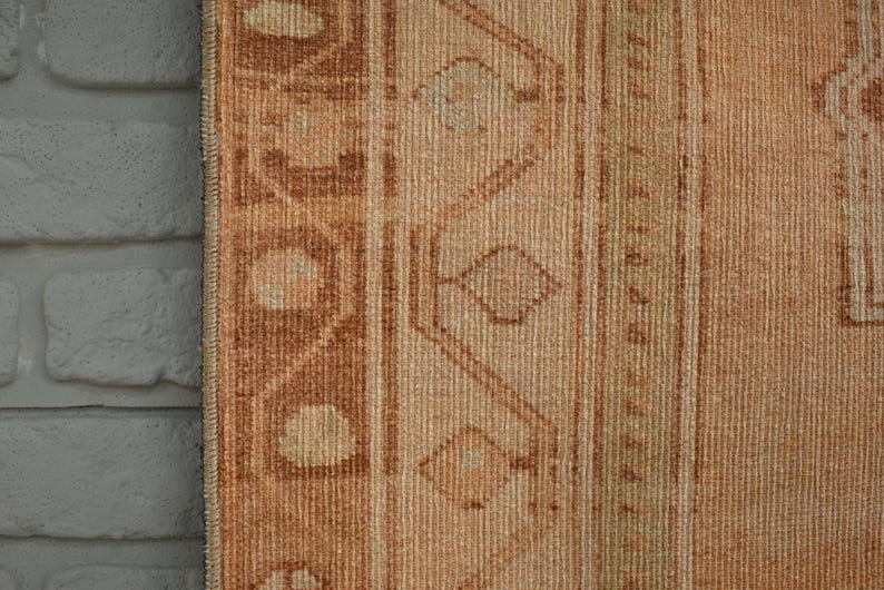 May include: Close-up of a section of a vintage rug with a brick wall on the left. The rug features a warm color palette of beige, brown, and tan. Geometric patterns and vertical stripes are visible.