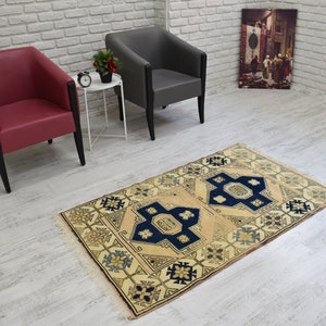 May include: A beige rug with a blue and yellow geometric pattern. The rug is on a white wood floor. Two chairs are in the background, one red and one gray.