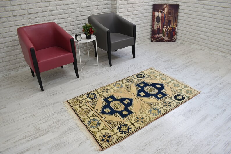 May include: A beige rug with a blue and yellow geometric pattern. The rug is on a white wood floor. Two chairs are in the background, one red and one gray.