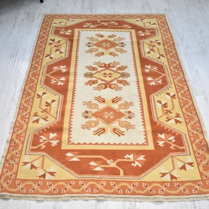 Puede incluir: Una alfombra turca tradicional con fondo crema y un patr&oacute;n geom&eacute;trico rojo, naranja y amarillo. La alfombra tiene un borde con flecos.