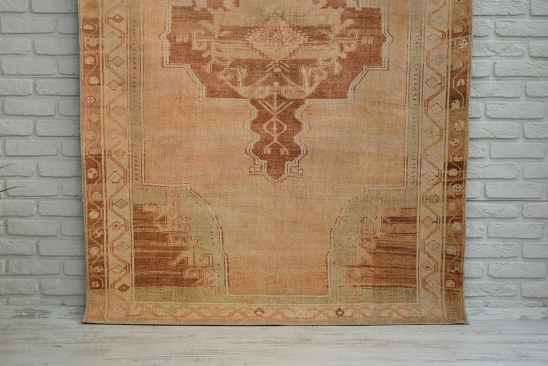 May include: A vintage-style rug with a central medallion design in shades of rust and beige. The rug features intricate border patterns and a distressed appearance, suggesting age and use. The rug is displayed against a white brick wall.
