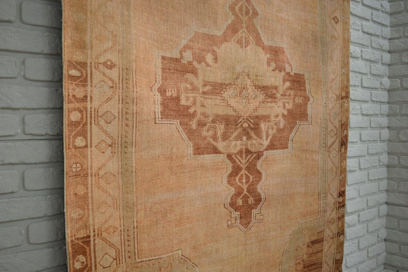 May include: A vintage rug with a central geometric design in shades of brown and beige. The rug has a distressed appearance, with faded colors and visible wear. The border features a repeating pattern, adding to the rug's traditional aesthetic.