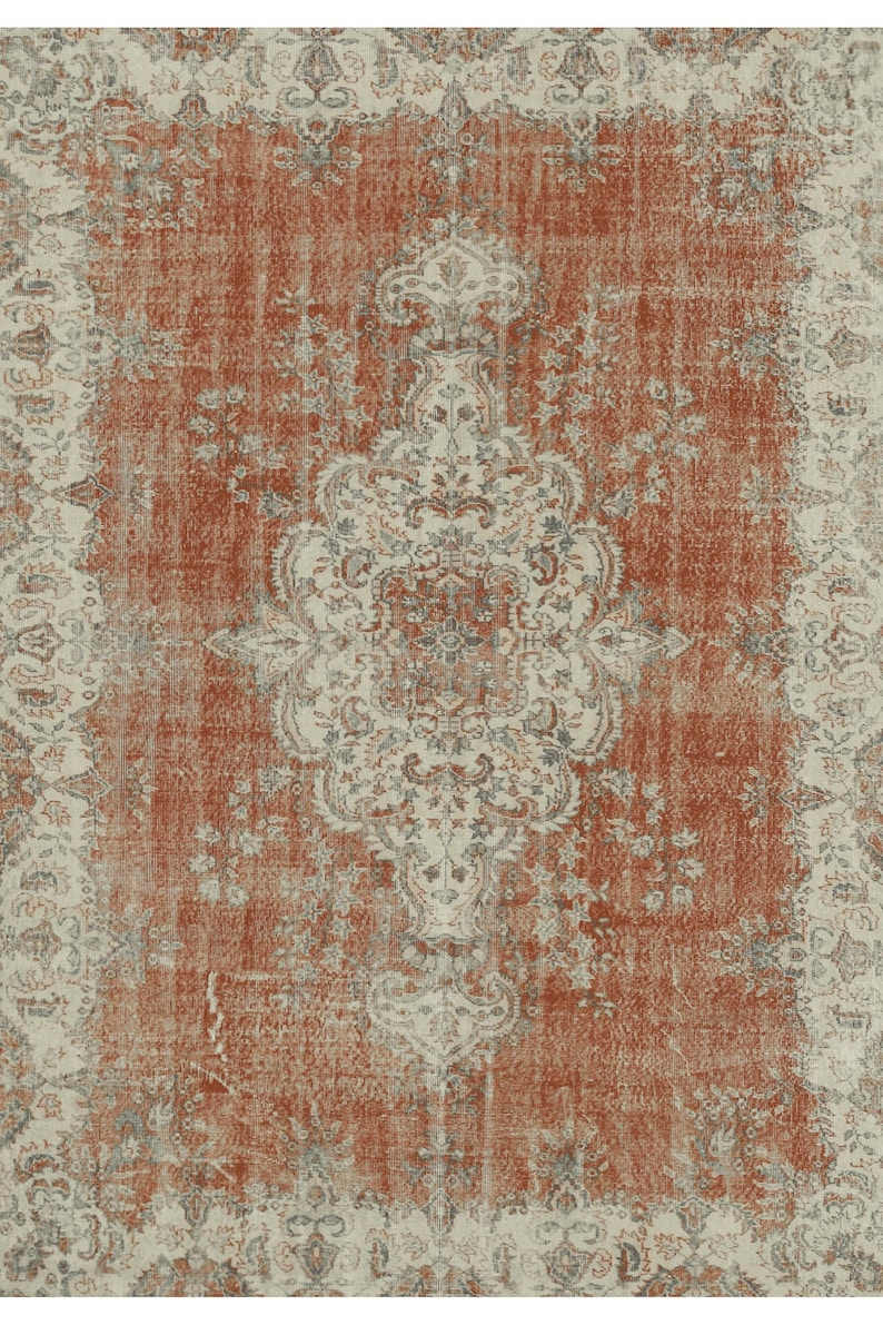 Puede incluir: Una alfombra de estilo vintage con un aspecto desgastado y descolorido. La alfombra presenta un medall&oacute;n central con intrincados patrones florales en tonos crema, gris y azul. El fondo es de un color naranja oxidado c&aacute;lido.