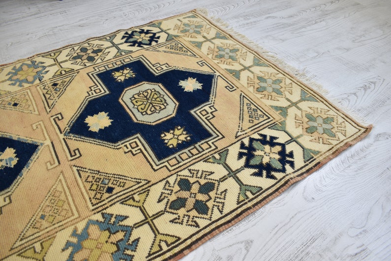 May include: A vintage rug with a cream background and intricate blue, green, and yellow geometric patterns. The rug has a fringed edge.