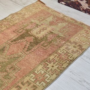 Puede incluir: Una alfombra turca vintage con fondo rosa y beige y un patr&oacute;n geom&eacute;trico verde. La alfombra tiene un aspecto desgastado y es perfecta para a&ntilde;adir un toque de estilo bohemio a tu hogar.