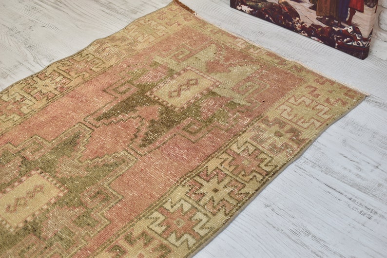 Puede incluir: Una alfombra turca vintage con fondo rosa y beige y un patr&oacute;n geom&eacute;trico verde. La alfombra tiene un aspecto desgastado y es perfecta para a&ntilde;adir un toque de estilo bohemio a tu hogar.