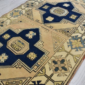 May include: A vintage Turkish rug with a beige background and intricate blue, green, and yellow geometric patterns. The rug features a central medallion with a repeating design.