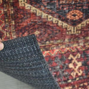 Op de afbeelding: Detail van een patroonkleed met een donkergrijze achterkant. Het kleed heeft een traditioneel ontwerp met rode, zwarte en beige kleuren. Een hand tilt een hoek van het kleed op en toont het rugmateriaal.