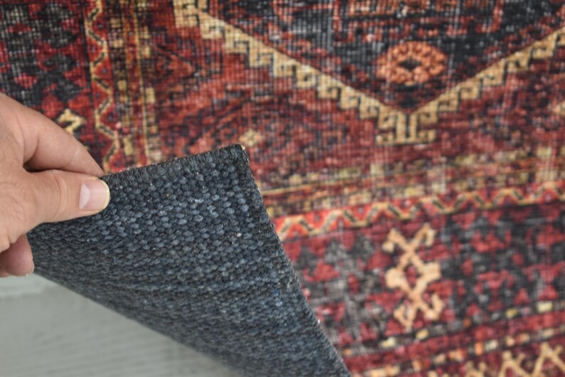 Op de afbeelding: Detail van een patroonkleed met een donkergrijze achterkant. Het kleed heeft een traditioneel ontwerp met rode, zwarte en beige kleuren. Een hand tilt een hoek van het kleed op en toont het rugmateriaal.