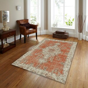Puede incluir: Una alfombra de estilo vintage desgastada con un fondo crema y un gran patr&oacute;n floral rojo-naranja intrincado. La alfombra est&aacute; colocada sobre un suelo de madera en una habitaci&oacute;n con un sill&oacute;n de cuero marr&oacute;n y una mesa auxiliar de madera.