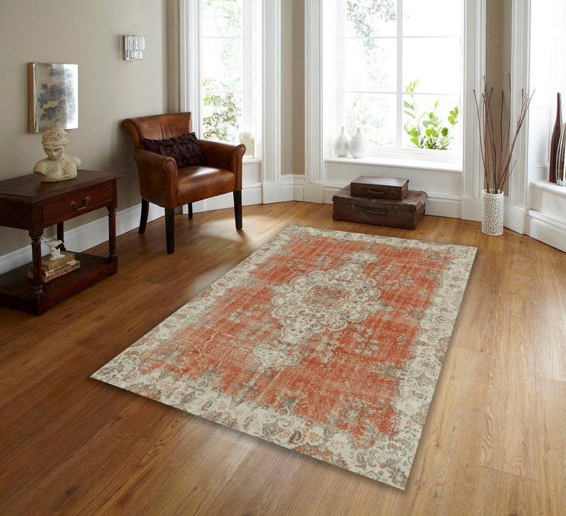 Puede incluir: Una alfombra de estilo vintage desgastada con un fondo crema y un gran patr&oacute;n floral rojo-naranja intrincado. La alfombra est&aacute; colocada sobre un suelo de madera en una habitaci&oacute;n con un sill&oacute;n de cuero marr&oacute;n y una mesa auxiliar de madera.