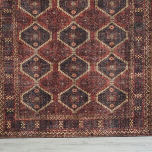 Op de afbeelding: Een rechthoekig tapijt met een geometrisch patroon. Het tapijt heeft een herhalend ontwerp van ruitvormen in tinten rood, bruin en beige. Het tapijt heeft een versleten, vintage uitstraling, met een gedetailleerde rand.