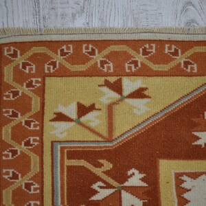 Puede incluir: Primer plano de una alfombra tejida con un patr&oacute;n geom&eacute;trico en tonos naranja, amarillo y blanco. La alfombra tiene un borde con flecos.