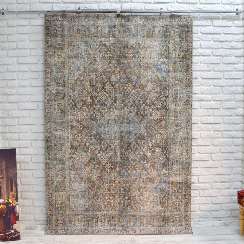 May include: A vintage-style rug with a brown, blue, and beige color scheme. The rug has a geometric pattern with intricate details. The rug is hanging on a white brick wall.